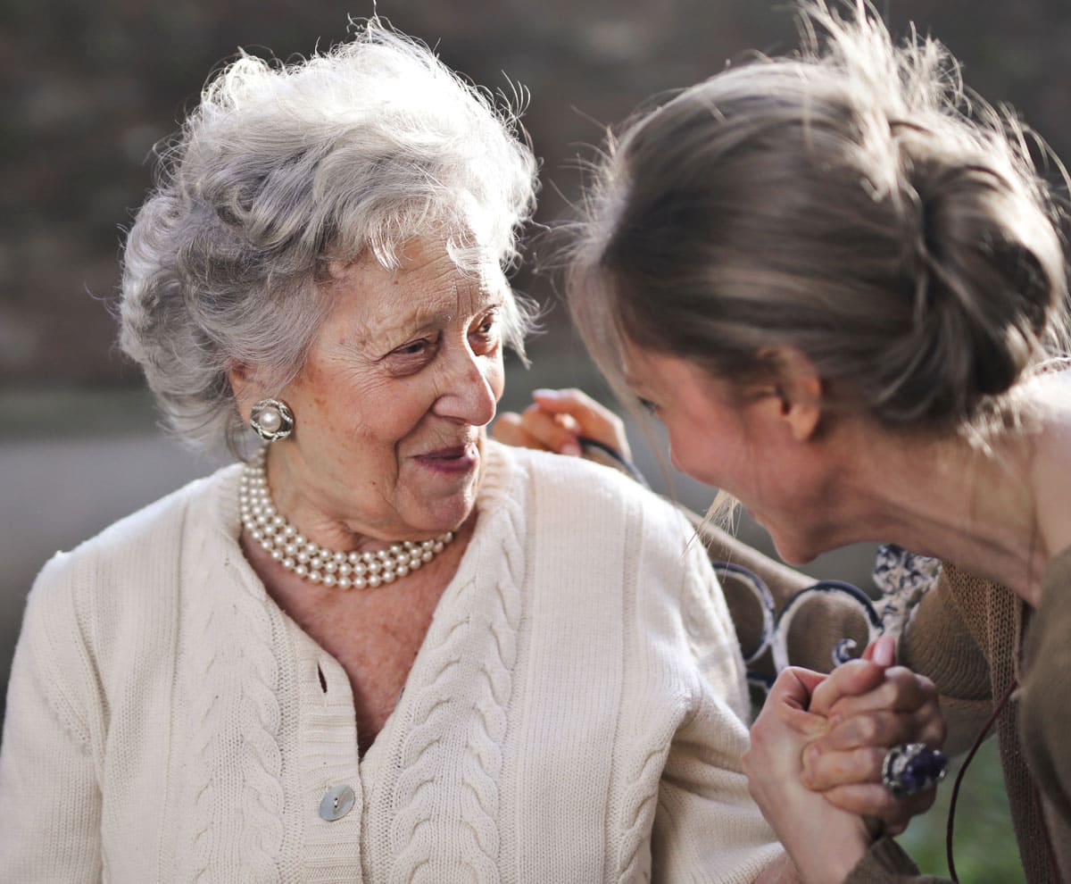 Una señora mayor y su cuidadora conversan sonrientes en un parque