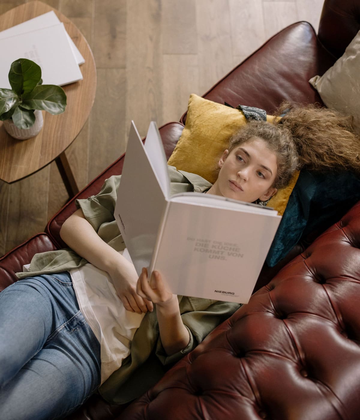 Una chica descansa tumbada en un sofá con la cabeza sobre unos cojines de colores mientras lee un libro